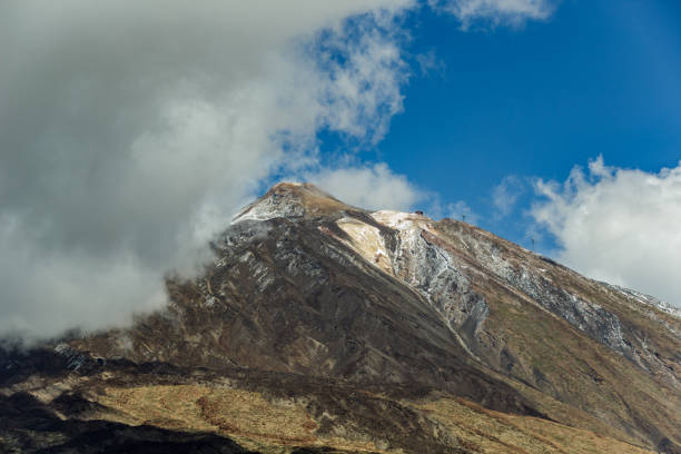 Mountain Teide with white snow spots, partly covered by the clouds. Bright blue sky. Teide National Park, Tenerife, Canary Islands, Spain.