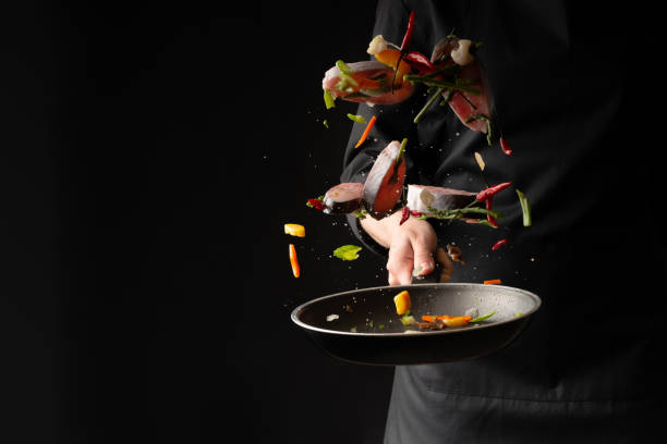 Chef cook fry fish with vegetables on a griddle on a black background. horizontal photo. sea food. healthy food. oriental cuisine, baner Chef cook fry fish with vegetables on a griddle on a black background. horizontal photo. sea food. healthy food. oriental cuisine, baner silver service stock pictures, royalty-free photos & images