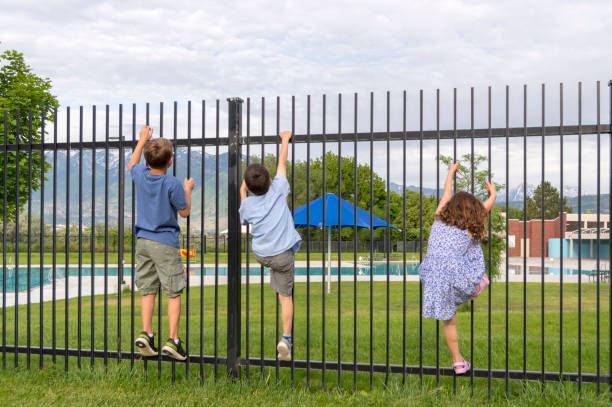 Three Children Climbing a Fence Guarding a Public Pool This is a group of children looking to scale the protective fence around a pool in a park on a summer day. The group consists of two boys and girl. There does not appear to be much chance of them making it over the fence. pool gate stock pictures, royalty-free photos & images