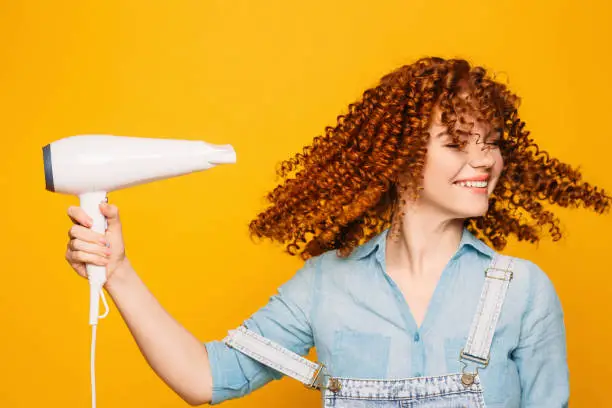 curly red-haired woman using hair dryer on yellow background. Making perfect curls