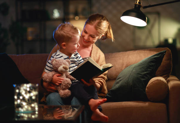 la famiglia prima di andare a letto la madre legge al libro del figlio vicino a una lampada la sera - bedtime stories foto e immagini stock