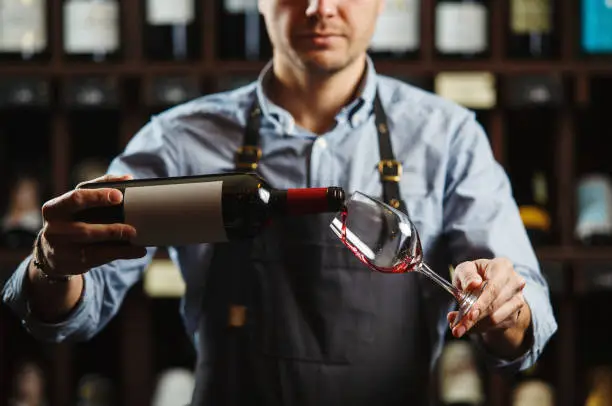 Male sommelier pouring red wine into long-stemmed wineglasses. Waiter with bottle of alcohol beverage. Bartender at bar counter pour elite drink into long-stemmed glass