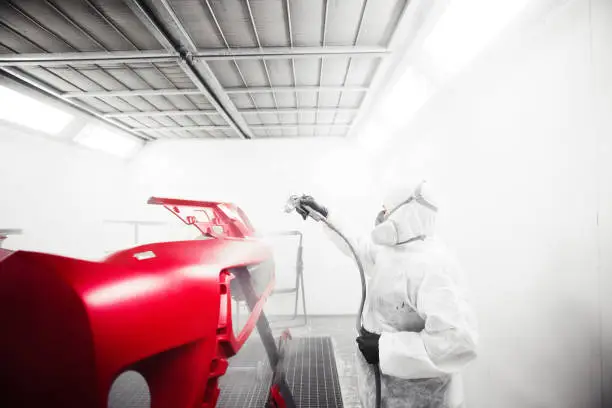 Male worker sprays varnish with spray gun on car bumper after painting. Auto repair garage.