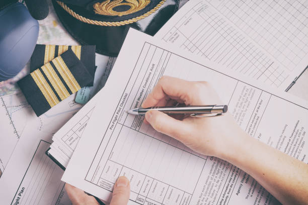 Close up of an airplane pilot hand filling in an pre-fligh checklist and holding weather forecast with equipment including hat, epaulettes and other documents in background