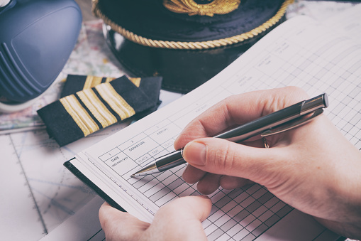 Close up of an airplane pilot hand filling in an pre-fligh checklist and holding weather forecast with equipment including hat, epaulettes and other documents in background
