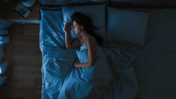 美麗的年輕女子睡在床在臥室在晚上的  頂視圖。藍色夜色與冷弱燈柱燈透過視窗閃耀。 - 睡覺 個照片及圖片檔