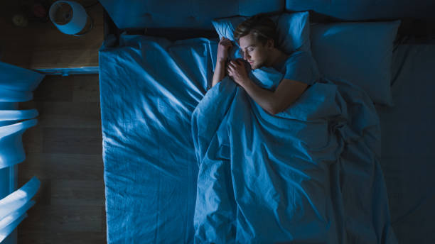 vista dall'alto del bel giovane che dorme pigramente su un letto nella sua camera da letto di notte. colori notturni blu con luce lampione debole fredda che brilla attraverso la finestra. - men young adult human head blond hair foto e immagini stock