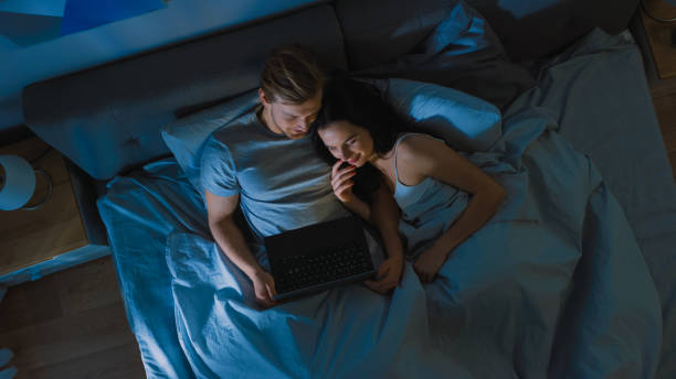 cute young couple in bed at night using laptop, watching series, reading social media, browsing through internet, talking and having fun. top view camera shot - lying down women laptop freedom imagens e fotografias de stock