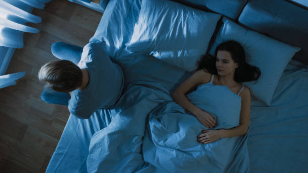 déprimé et stressé jeune homme s'assied sur le bord du lit la nuit, souffrant d'insomnie en raison de problèmes sexuels. sa jeune femme se trouve à côté de lui sentant l'empathie. - dark edge photos et images de collection