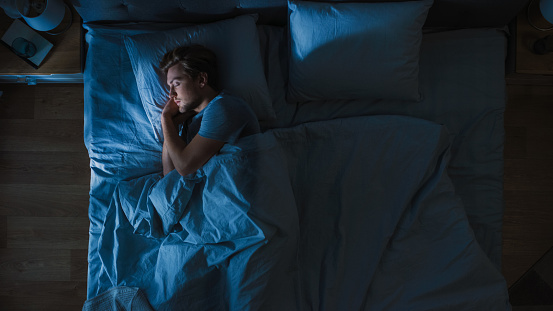 Vista superior del joven guapo durmiendo acogedoramente en una cama en su dormitorio por la noche. Colores azules nocturnos con luz de farola débil fría que brilla a través de la ventana. photo