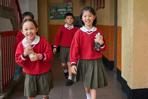 School children holding water bottles while walking in lobby. Happy students are taking break in campus. They are wearing school uniform.