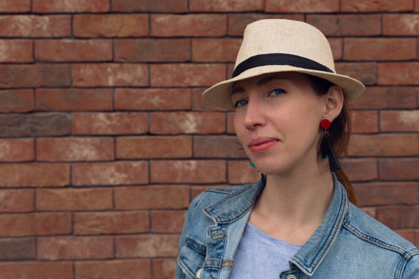 istock Beautiful young woman in hat is looking at camera confidently and smiling while standing against white brick wall 1156697365