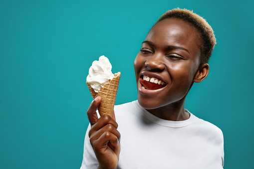 View from side of happy young girl looking at delicious ice cream on blue isolated background in studio. Cheerful in white shirt keeping waffle and smiling. Concept of satisfaction and summer.