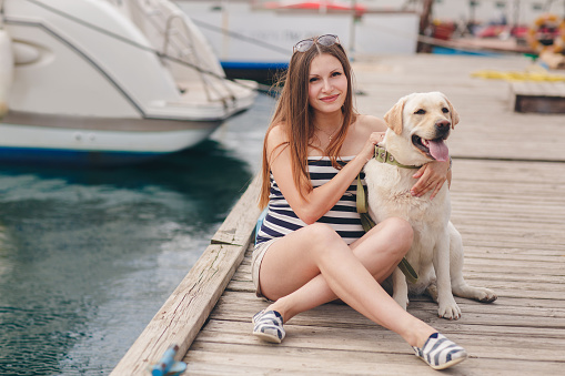 Beautiful young pregnant woman,a brunette with long straight hair and brown eyes,wears a gold neck chain and cross,sits on the dock near the sea on the backdrop of the yacht with his dog breed white Labrador