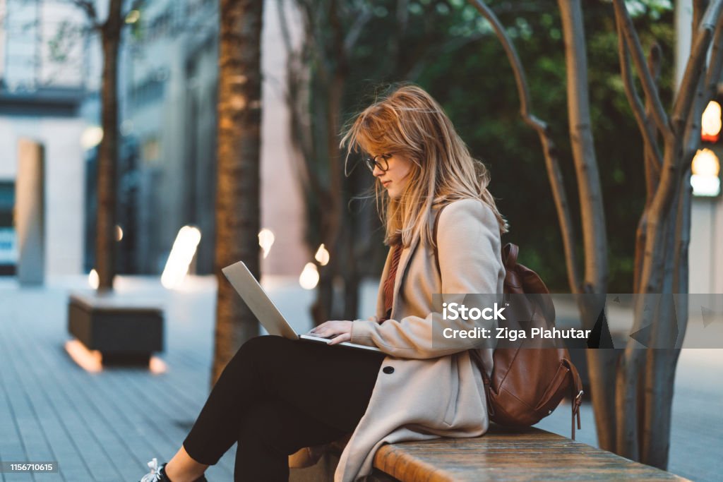 Woman using laptop on a bench Young businesswoman outside, walking, having a phone conversation, working, having a video call. Student sitting on a bench in a park while using laptop and mobile phone at dusk. Outdoors Stock Photo