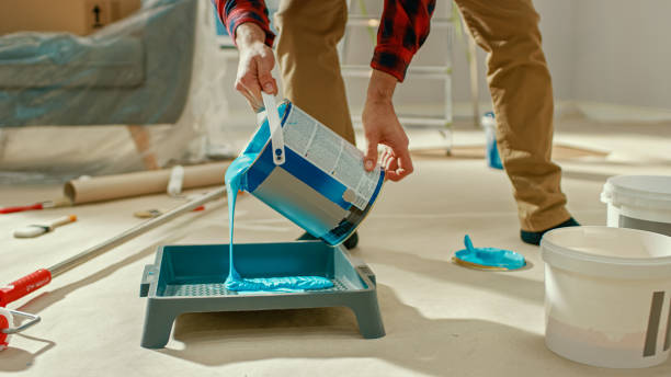 o homem novo na calças de brim de brown e na camisa verific vermelha pega uma lata da luz-pintura azul e derrama-a em uma bandeja. renovações de quarto em casa. - blue construction built structure indoors - fotografias e filmes do acervo