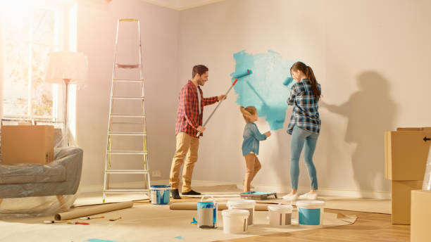 hermosa familia joven están mostrando cómo pintar paredes a su adorable hija pequeña. pintan con rodillos que están cubiertos de pintura azul claro. disparo con destello de sol en la habitación renovada en casa. - female house painter home decorator paintbrush fotografías e imágenes de stock