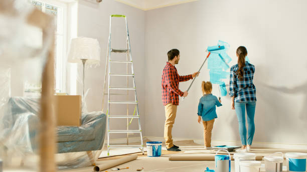 美しい若い家族は、彼らの愛らしい小さな娘に壁をペイントする方法を示しています。彼らはライトブルーの塗料で覆われているローラーでペイントします。自宅の部屋の改装。 - female house painter home decorator paintbrush ストックフォトと画像