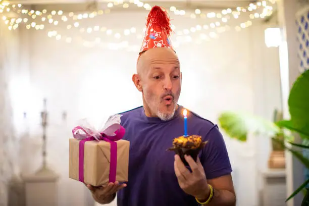 Man blows out a candle on the cake, holding a present in his hand - birthday celebration concept