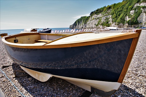 A unique study of empty, abandoned boats on an equally empty beach at the English Channel resort of Beer in South Devon, near Exeter, part of the Jurassic Coast World Heritage Site. The images were all taken on early summer's sunny day when the beach would normally be teeming with people but there is no-one to be seen. The boats all vary in size, shape and purpose, many are covered in tarpaulins but all are neatly stranded on the beach in rows as if waiting for passengers. The emptiness of the boats coupled with the lack of any people lends a surreal atmosphere to the images almost like the Marie Celeste. The sunny, bright day adds to the colourful imagery and invites the viewer to explore the beach. This 8th and final image in the series is of a very smart and well-maintained single rowing or fishing boat with a high level of gloss navy blue paint on the hull and a highly polished and varnished, natural wood interior. The rows of empty red-striped deckchairs in the distance and the reflection of the pebbles on the beach in the glossy hull paint of the boat indicate recent abandonment, almost as if the crowds were running away from radiation or some other invisible force.