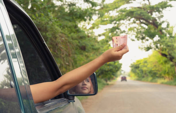 Young female driver paying a road toll service charge from a car Young Asian woman holding out paper money through a car window to pay a toll fee - Tanned millennial female holding Thai currency banknote from a vehicle to pay a corruption bribe cash for cars stock pictures, royalty-free photos & images