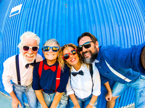 istock Four smiling people take selfie in front to a blue wall. Dresses  with colored bow tie and suspenders, smiling at camera. Emotion and love for two couples of parents 1156729567