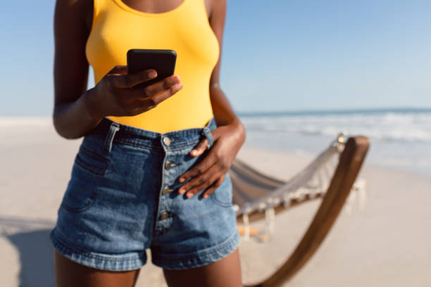 frau mit handy mit den daumen in gürtelschlaufen am strand - beach shorts stock-fotos und bilder