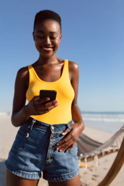 istock Woman using mobile phone with her thumbs in belt loops on the beach 1156732053
