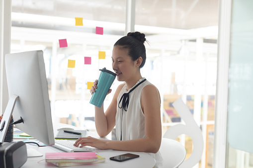 Side view of Asian female graphic designer drinking hot beverage while working on computer at desk in a modern office