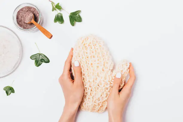 Woman's hands holding natural massage mitten washcloth next to coffee body scrub and bowl of sea salt on white background, top view.