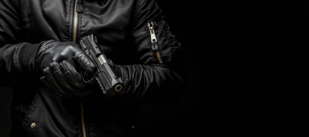 un uomo con una giacca nera e guanti neri in possesso di una pistola su uno sfondo scuro - gun gunman handgun men foto e immagini stock