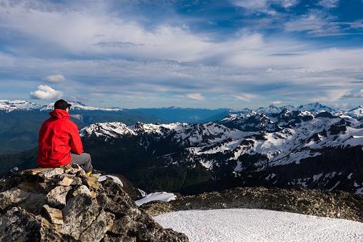 Rear view of hiker looking at snow covered mountains. Mature backpacker is sitting on rocks against cloudy sky. He is wearing red hooded jacket.