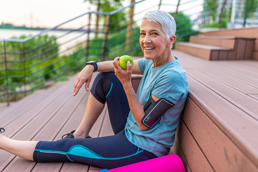 Sporty woman eating apple. Beautiful woman with gray hair in the early sixties relaxing after sport training. Healthy Age. Mature athletic woman eating an apple after sports training