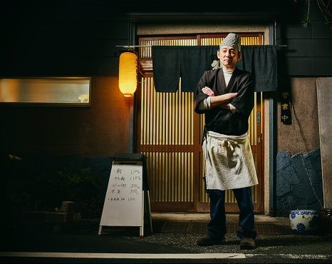A mature aged Japanese sushi chef standing in a front of his shop at night.