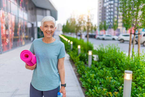 Gorgeous senior woman carrying a yoga mat outdoors and smiling Beautiful senior woman with a yoga mat. Pretty senior sportswoman holding fitness mat while standing outdoors. Portrait of mature woman with yoga mat yoga class photos stock pictures, royalty-free photos & images