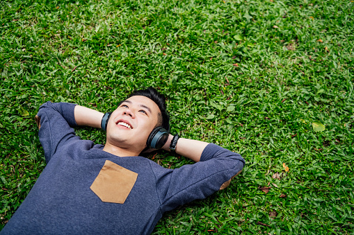 Happy young Chinese man enjoying music, lying with hands behind head on grass, happiness, contemplation, escapism