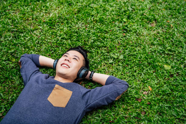 Young man lying on grass wearing headphones Happy young Chinese man enjoying music, lying with hands behind head on grass, happiness, contemplation, escapism asian man headphones stock pictures, royalty-free photos & images