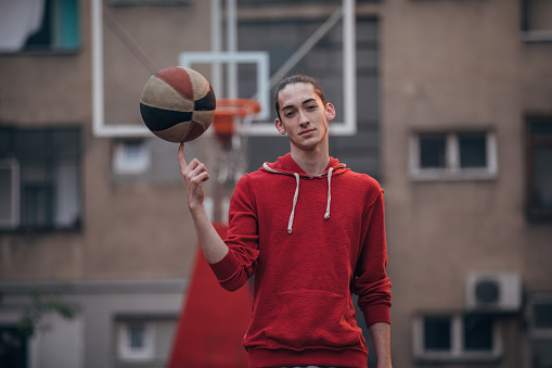 Handsome young man in red hooded sweatshirt standing on a basketball court and holding basketball on one finger.