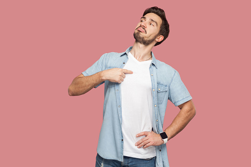 This is me. Portrait of proud haughty handsome bearded young man in blue casual style shirt standing, looking away and pointing himself. indoor studio shot, isolated on pink background.