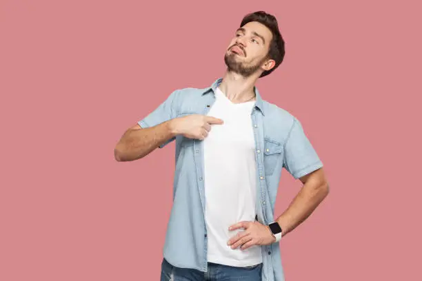 This is me. Portrait of proud haughty handsome bearded young man in blue casual style shirt standing, looking away and pointing himself. indoor studio shot, isolated on pink background.