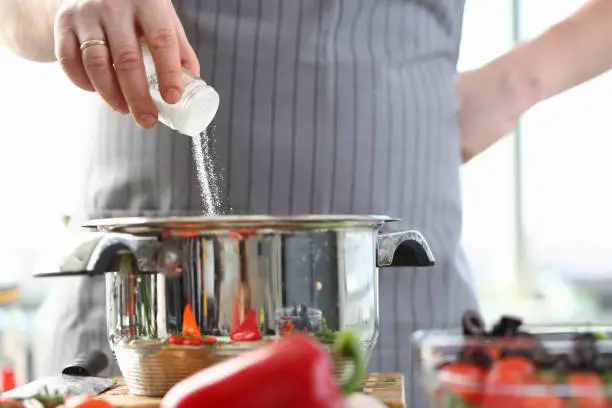 Culinary Chef Adding Saucepan White Sea Salt. Man in Apron Holding Spice Shaker in Hand. Male Fingers Putting Ingredient to Stainless Pan. Cooking in Kitchen at Home Horizontal Photography