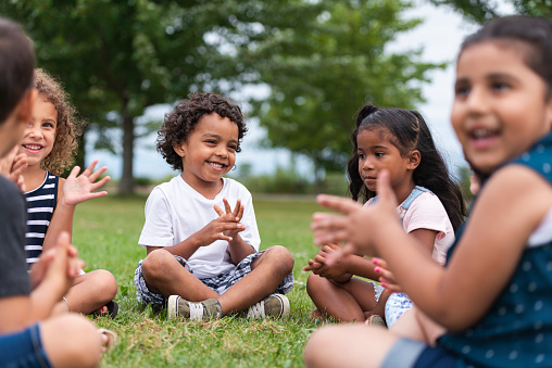 An adorable group of multi-ethnic children are outside sitting in the grass one sunny afternoon. They are clapping and smiling while sitting cross-legged and singing a song.