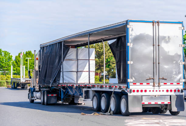 istock Big rig semi truck with open covered semi trailer unloading commercial cargo on warehouse parking lot 1156827220