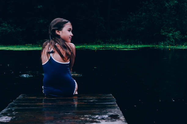 Girl sitting on pier looking back. Child swimming in lake. Spending time actively outdoors. Childhood memories. Having time outside in summer. Beautiful scenic background, backdrop