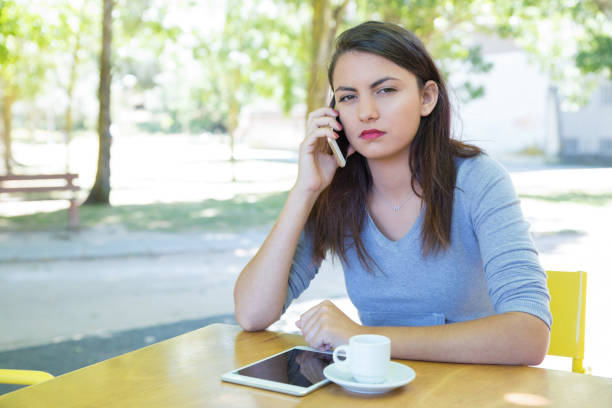 Serious pretty lady talking on phone at cafe table in park. Lady wearing hoodie, sitting and looking at camera with blurred green trees in background. Leisure and communication concept. Front view.