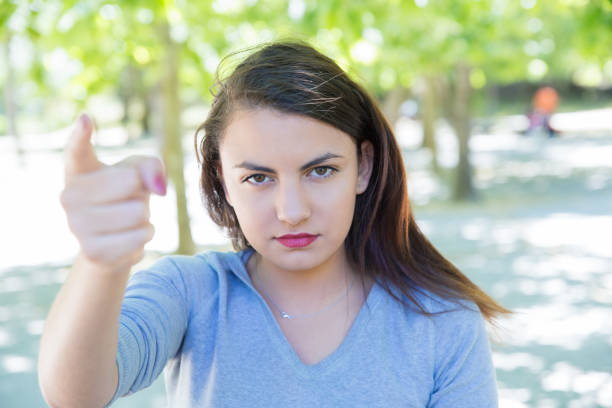 Serious pretty young woman accusing viewer in park. Beautiful lady looking and pointing at camera with blurred green trees in background. Accusation concept. Front view.