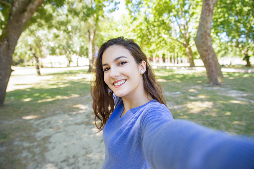 Happy young woman in blue sweater talking selfie in park. Carefree girl with wavy hair having fun alone in summer park. Environment concept