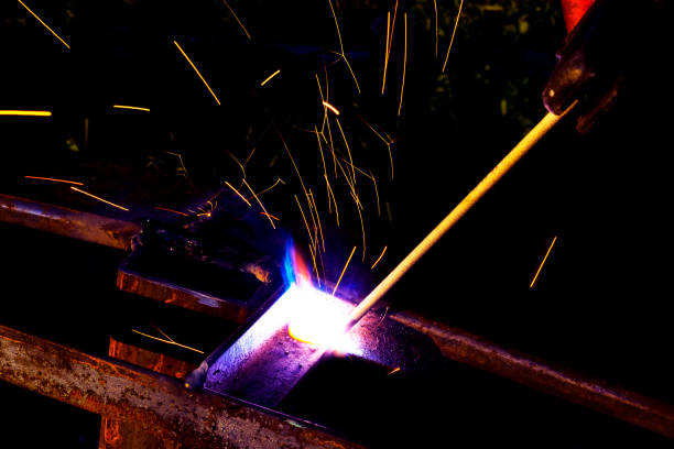 istock Welder worker performs jump welding. Man welder in protective gloves performs arc-welding process of metal structures. Flying sparks from the welding machine. Background construction site 1156872367
