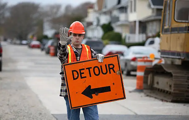 Road construction worker holding a detour sign and gesturing to stop.