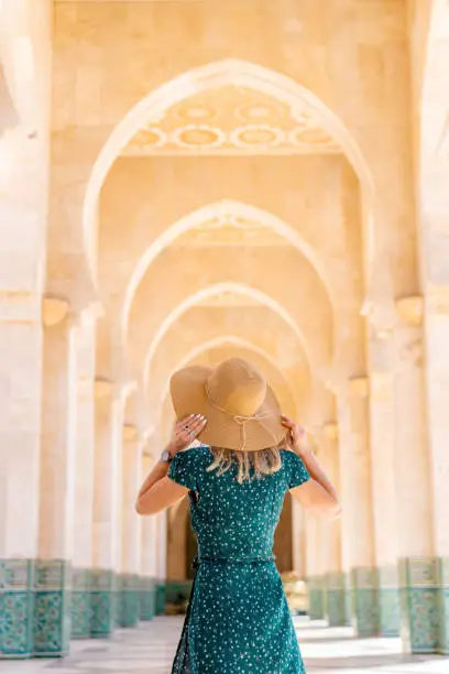 Young woman wearing sunglasses, green dress and wicker hat walking in ornate arch corridors of the Hassan II mosque.